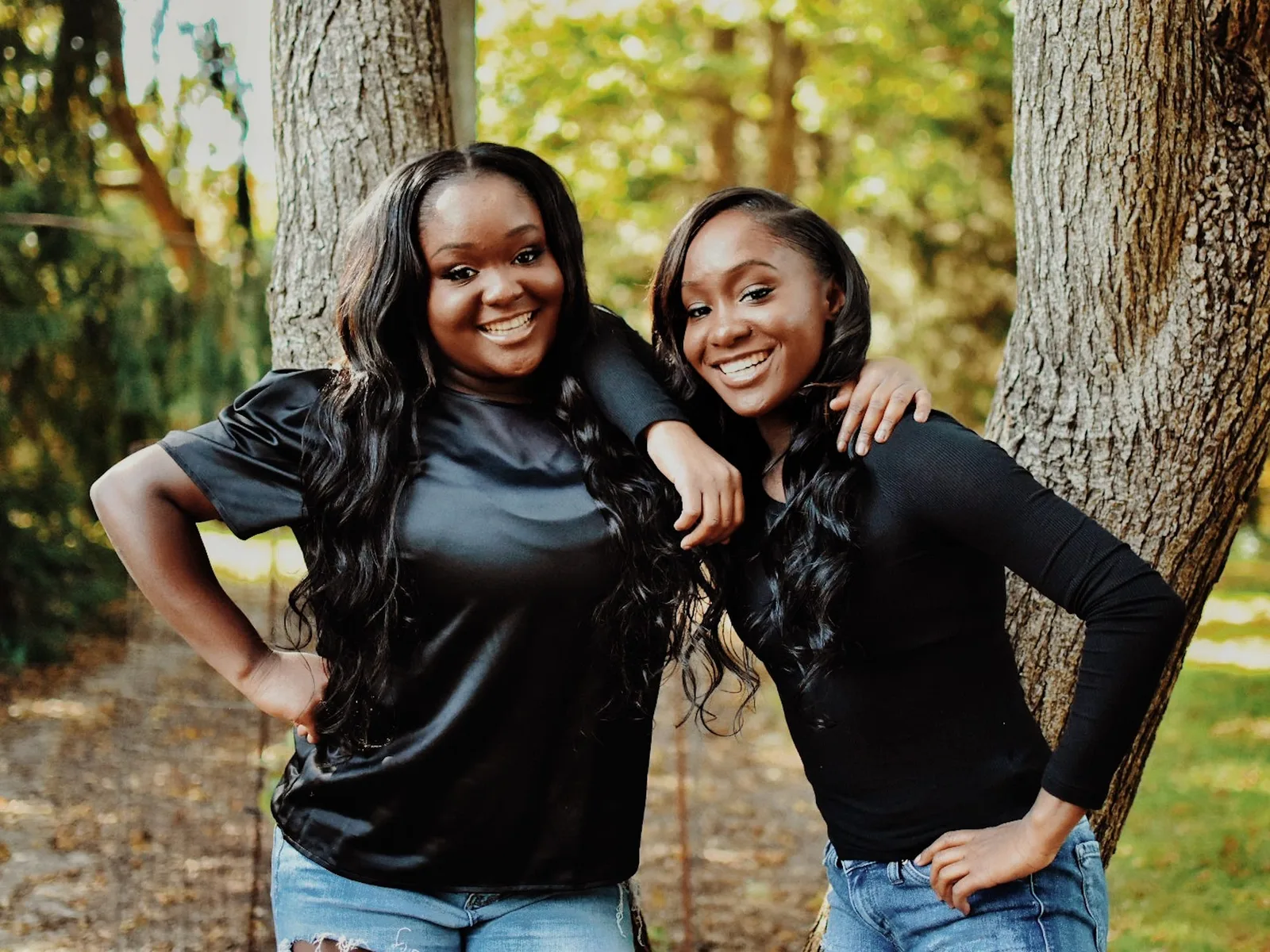 Two twin sisters standing arm in arm beside a tree, one heavier than the other