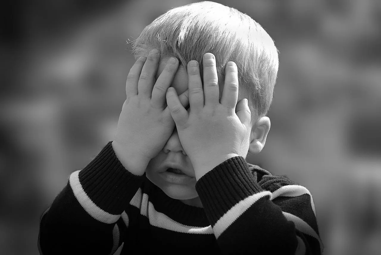 A black and white photograph of a boy covering his face with both hands