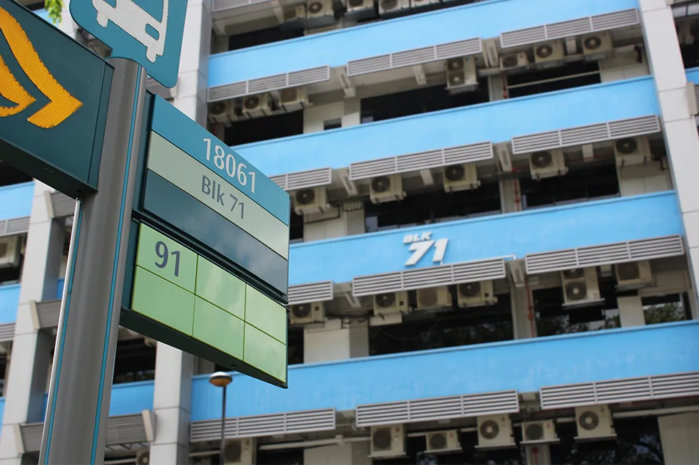 The blue and white BLK71 building, air-conditioning units along every floor, behind a bus stop sign reading 18061 Blk 71
