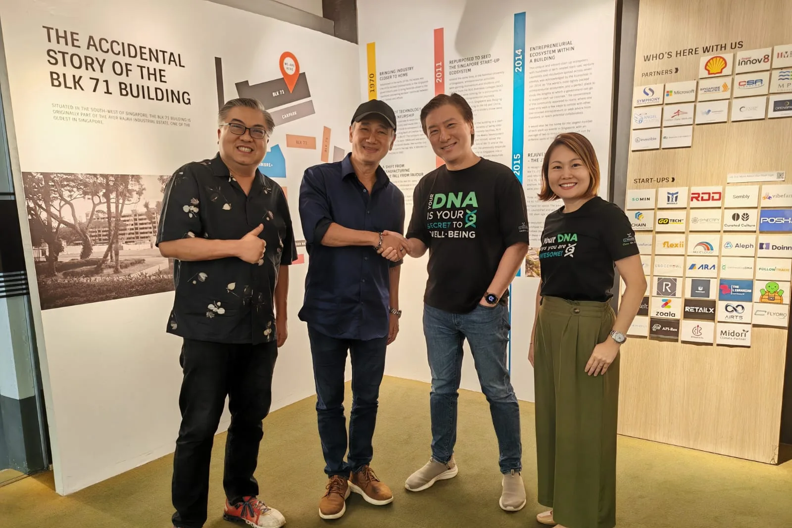 Gurmit Singh in a cap and navy shirt shaking hands with one of the ConnectingDNA team in a black DNA tee, a man in a patterned shirt giving a thumbs up and a woman in a ConnectingDNA tee beside them, in front of a wall headed The Accidental Story of the BLK 71 Building