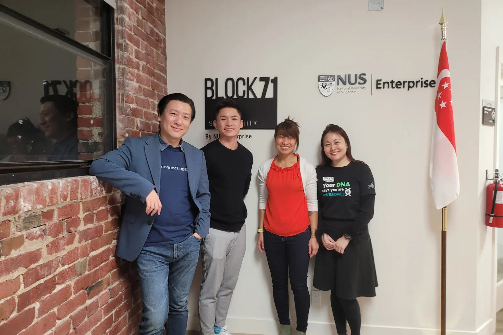 One of the ConnectingDNA team in a blue blazer over a ConnectingDNA T-shirt, leaning on a brick wall, with a man and two women beside him under the BLOCK71 Silicon Valley and NUS Enterprise signs and a Singapore flag