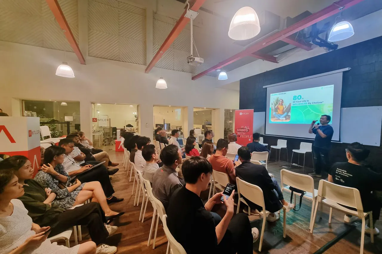 Cedric Lee holding a microphone beside a projected slide reading 80% of your life is influenced by choices, rows of seated people in white chairs facing him under a high ceiling with red beams, an Antler banner at the side