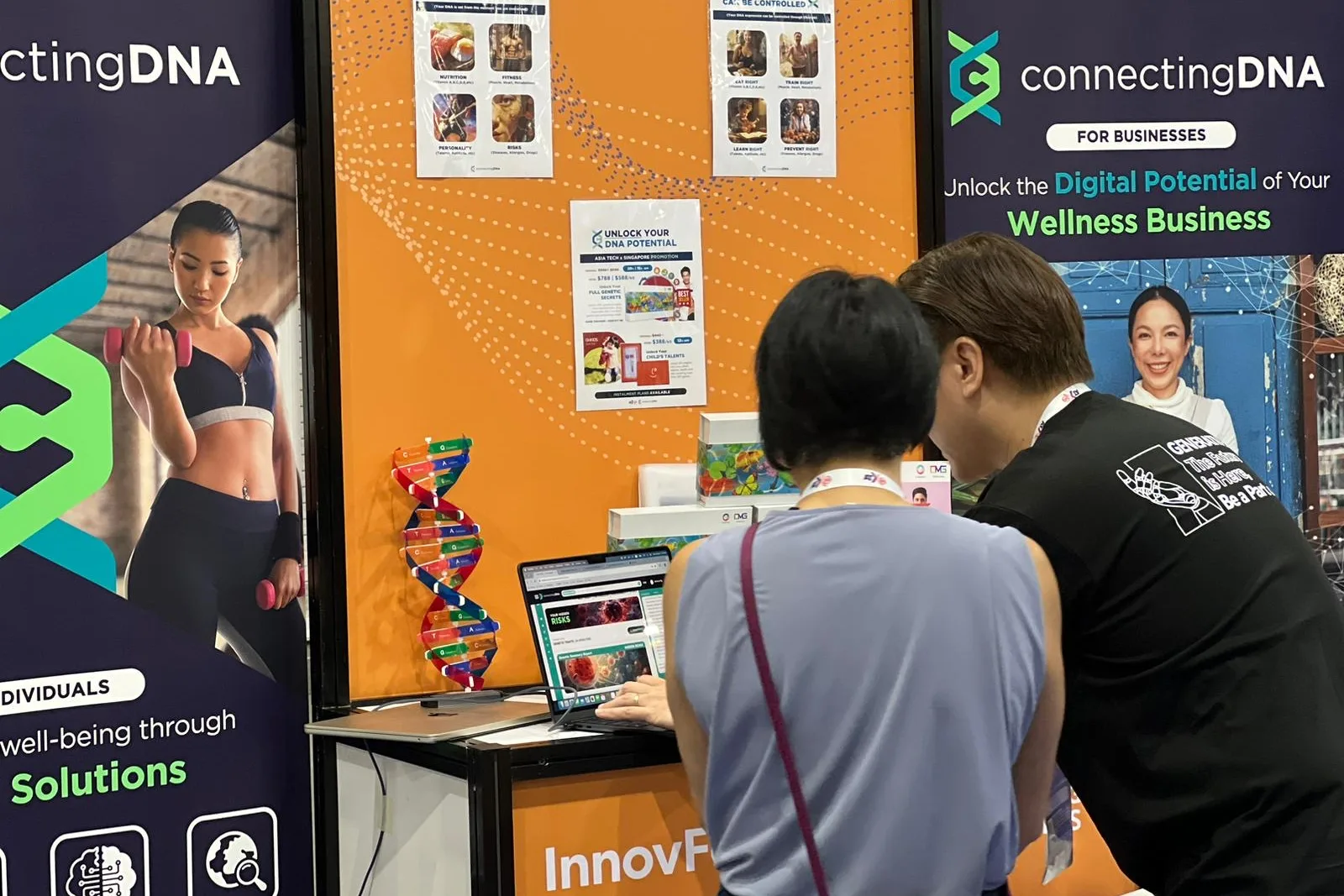 A visitor bent over a laptop at the ConnectingDNA booth with one of the team, the orange backdrop reading Unlock the Digital Potential of Your Wellness Business, an InnovFest sign on the counter