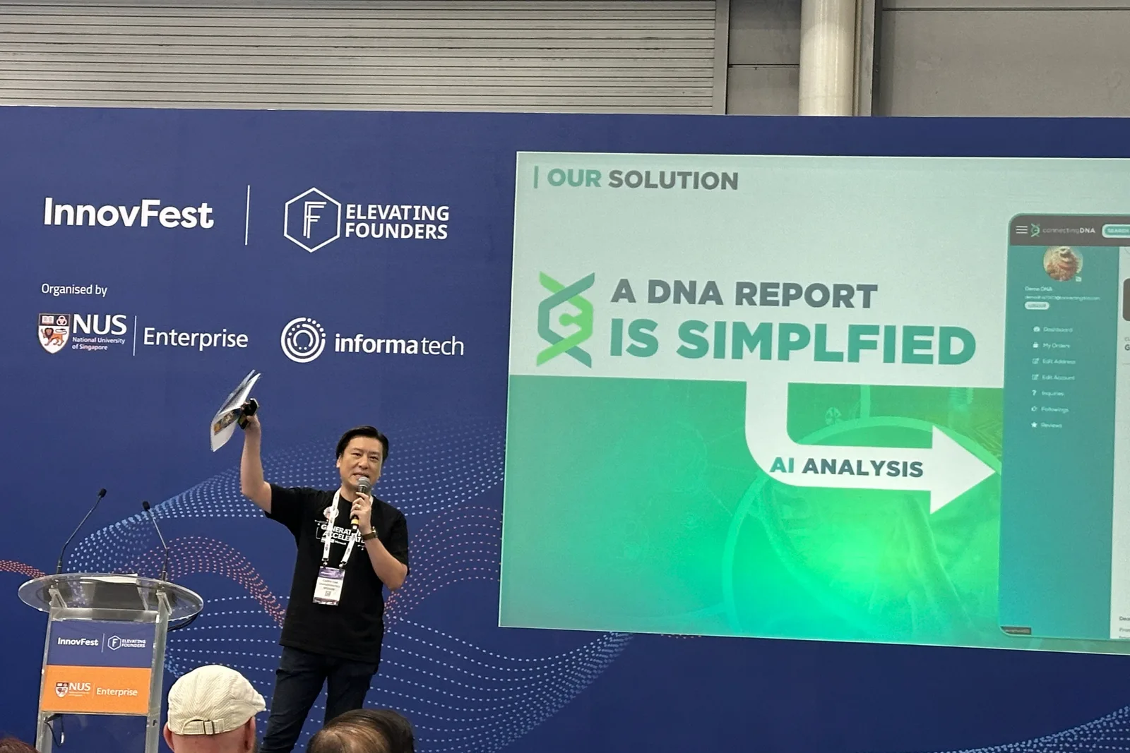 Cedric Lee on stage holding up a printed DNA report and speaking into a microphone, beside a screen reading Our Solution, A DNA Report Is Simplified, AI Analysis, with InnovFest and NUS Enterprise banners behind him