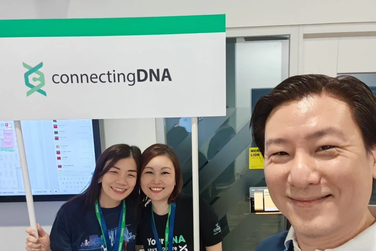 The ConnectingDNA team at their booth sign, one giving a thumbs up, two under it with a laptop and a card reading The World's First DNA-Wellness Marketplace