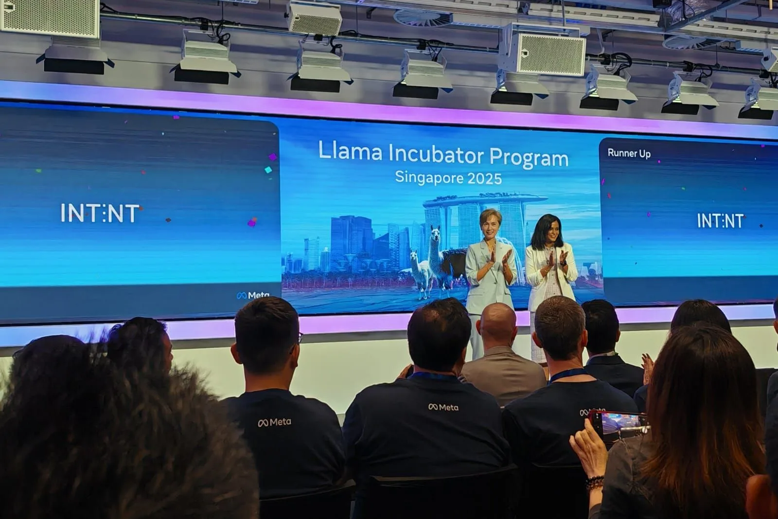 A stage screen reading Llama Incubator Program Singapore 2025, two women applauding on it and Runner Up beside it, seen over an audience in Meta shirts
