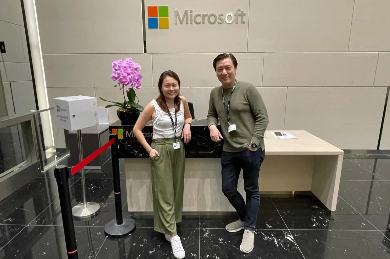 Cedric Lee and Ee Ting standing at a reception desk under a Microsoft sign, an orchid on the counter