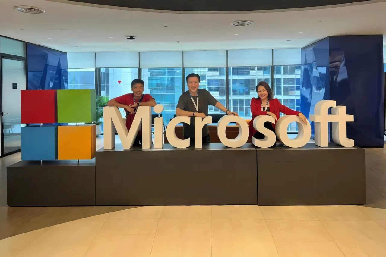 Three of the ConnectingDNA team leaning on the large white Microsoft letters in an office, the four-colour Microsoft logo at the left