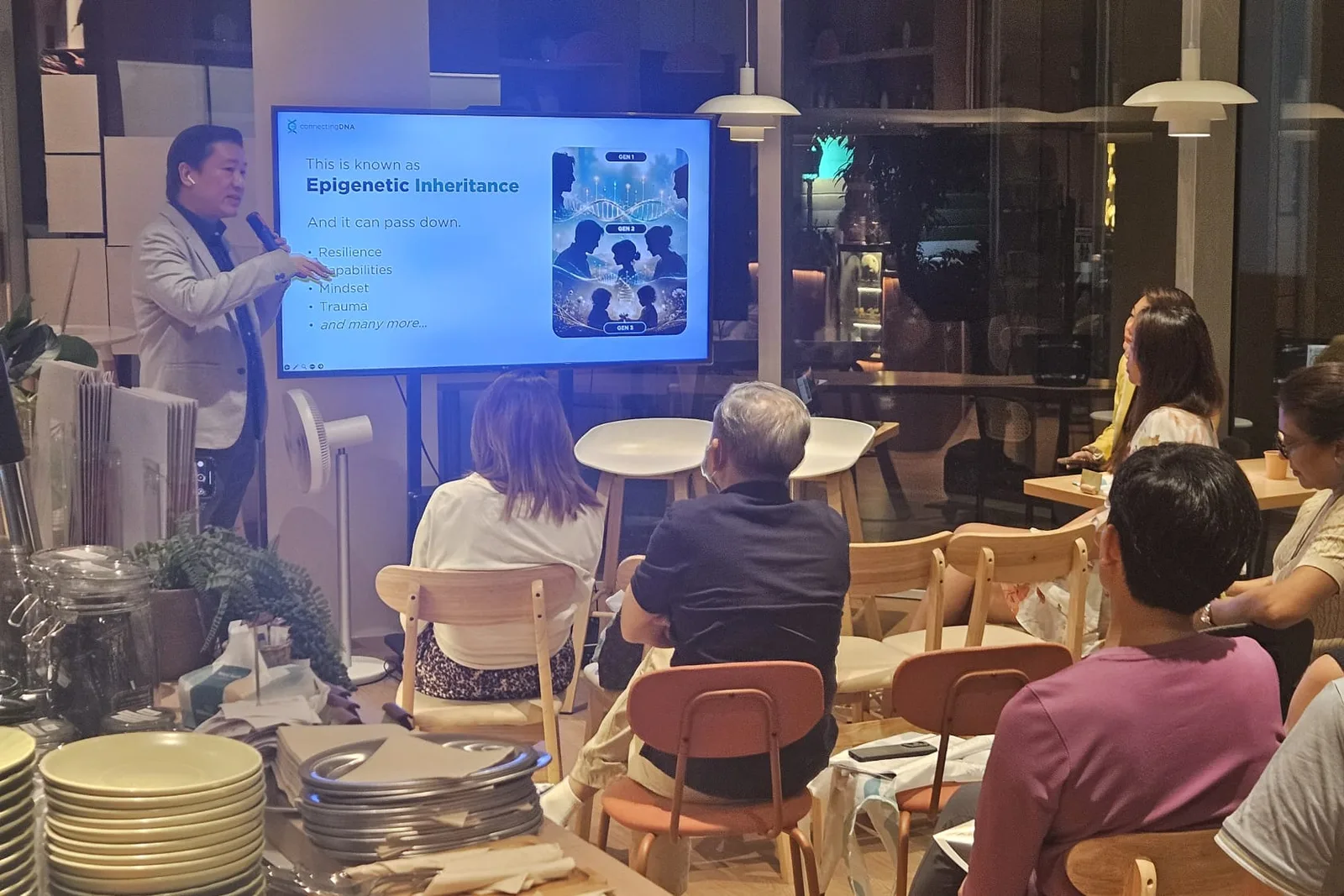 Cedric Lee in a grey blazer presenting beside a screen reading This is known as Epigenetic Inheritance, a seated audience at cafe tables in front of him