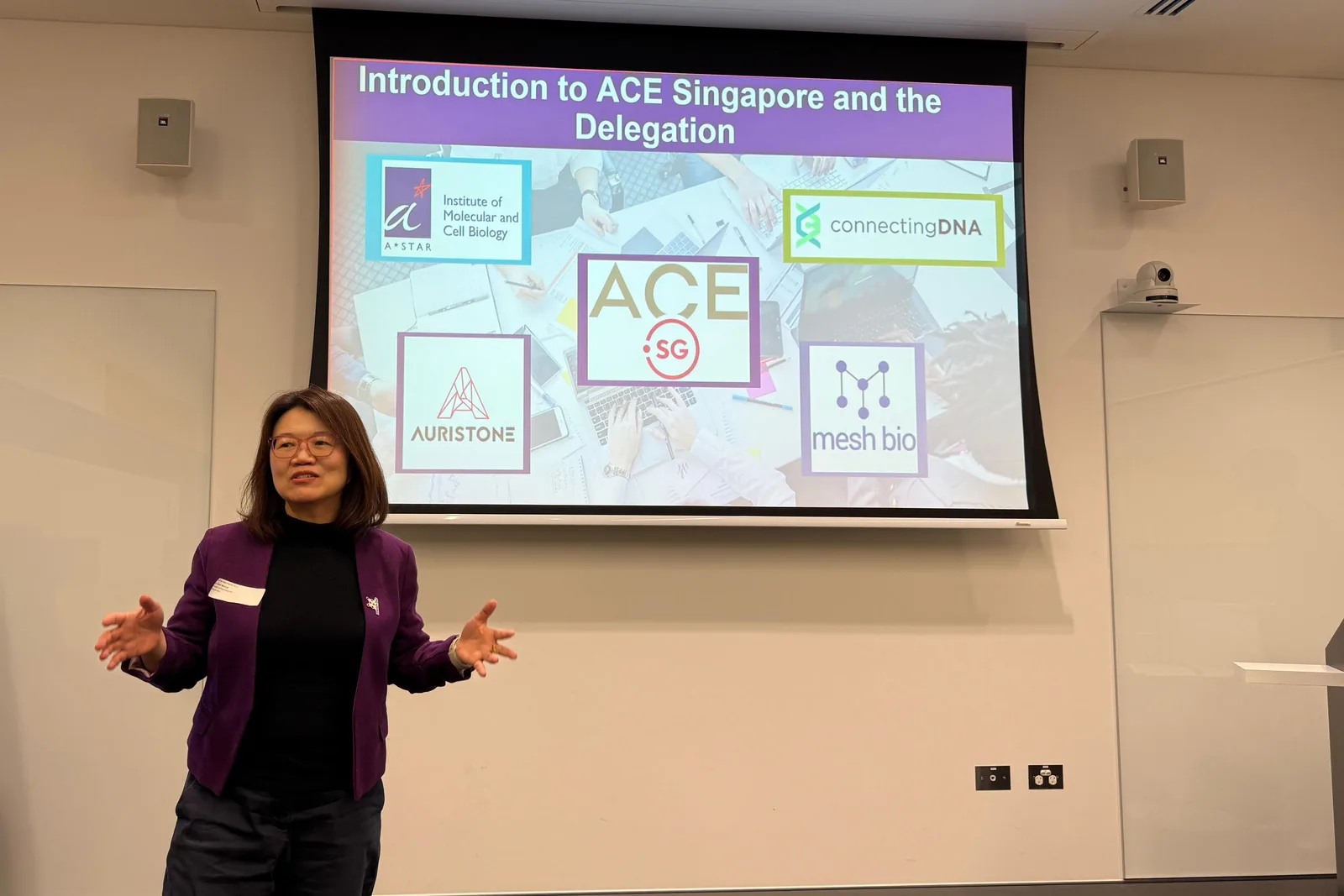 A woman speaking with her arms open before a screen headed Introduction to ACE Singapore and the Delegation, the ACE.SG, ConnectingDNA, Mesh Bio, Auristone and Institute of Molecular and Cell Biology logos on it