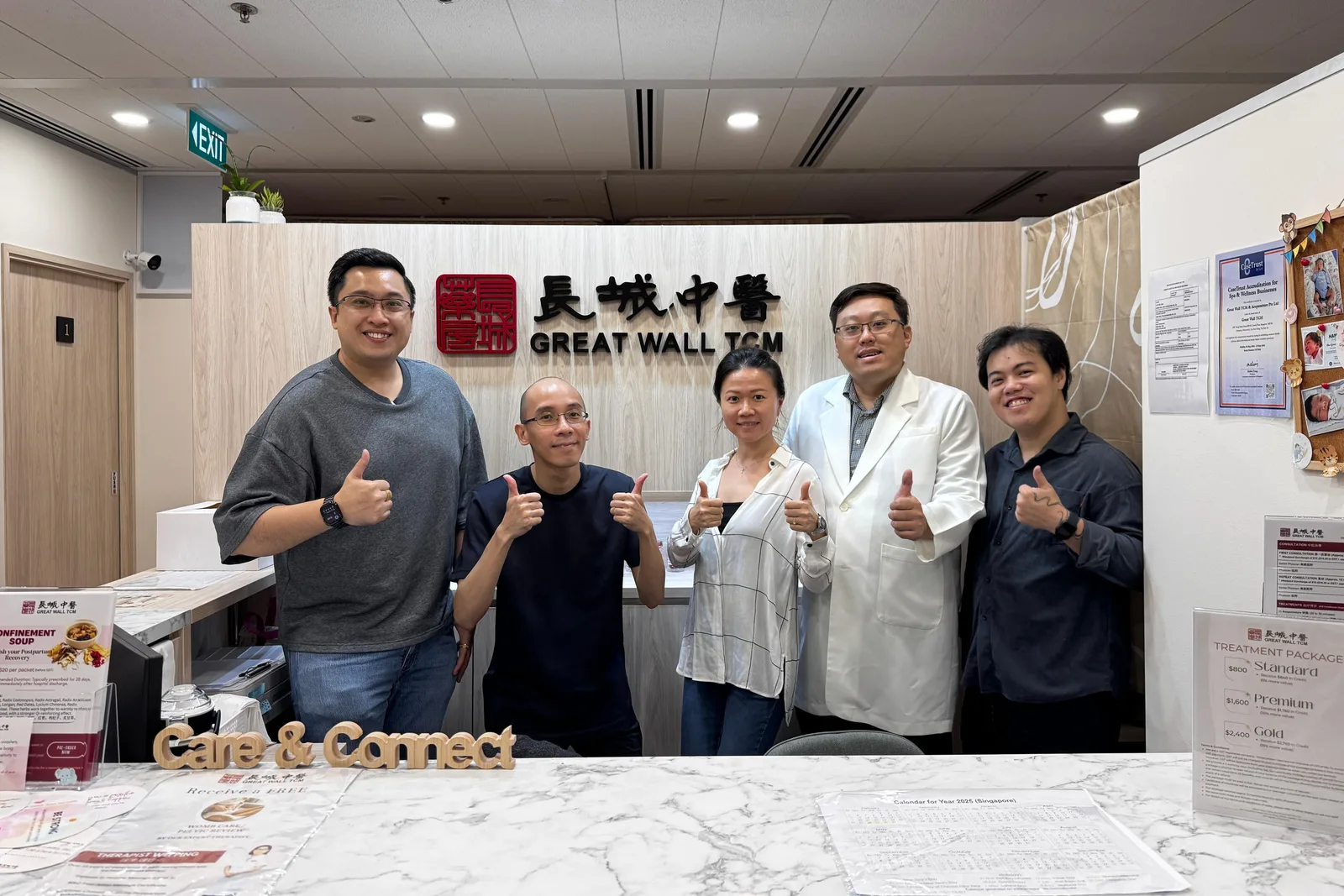 Six people giving a thumbs up behind a clinic's marble reception counter, two practitioners in white coats among them and a Great Wall TCM sign on the wall behind