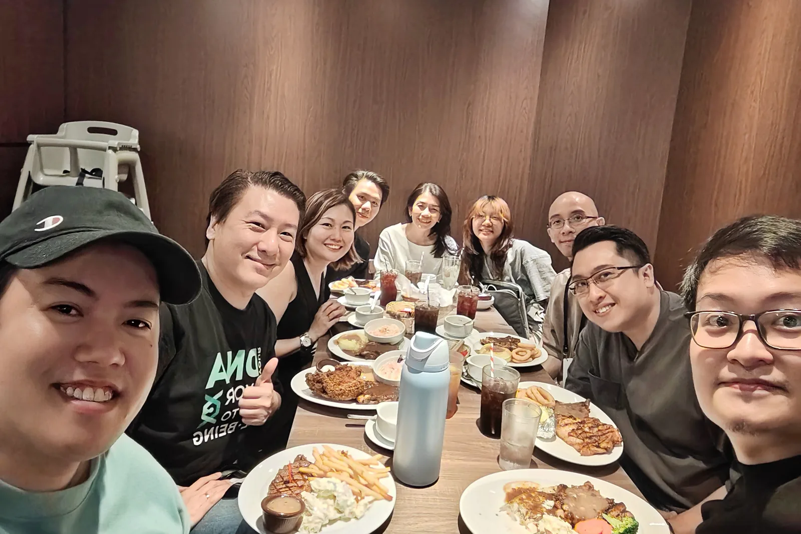 Ten of the ConnectingDNA team smiling for a selfie around a long restaurant table set with steaks, fries and iced drinks, one giving a thumbs up