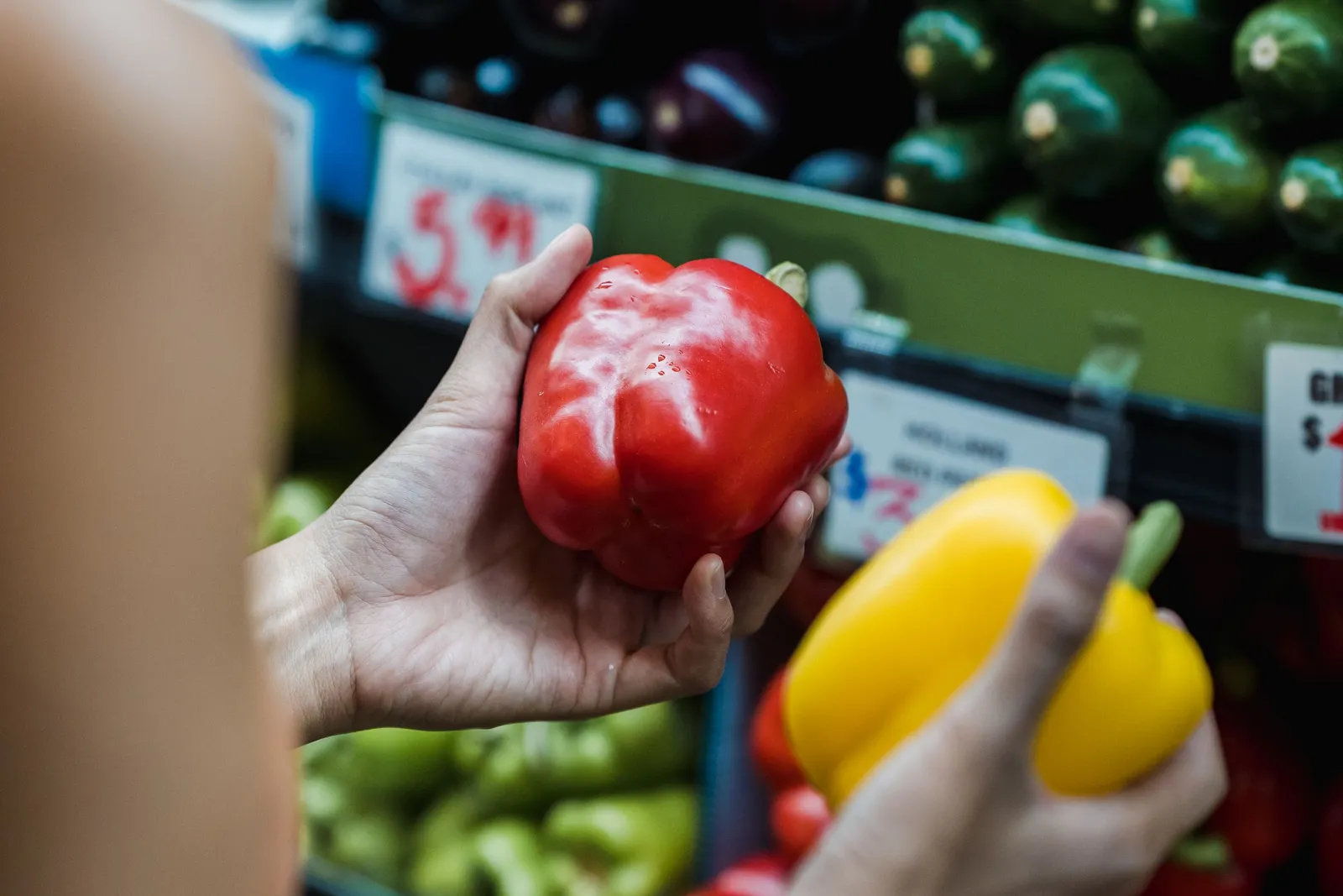A woman at a supermarket shelf holding a red pepper in one hand and a yellow one in the other