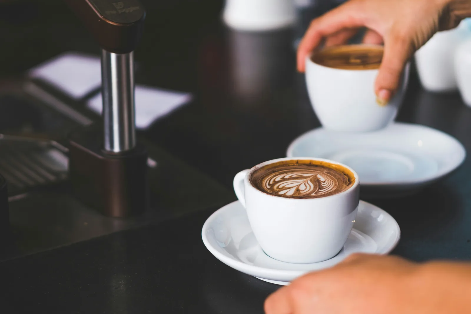 A cup of coffee with latte art set down on a dark counter, a second cup being lifted behind it