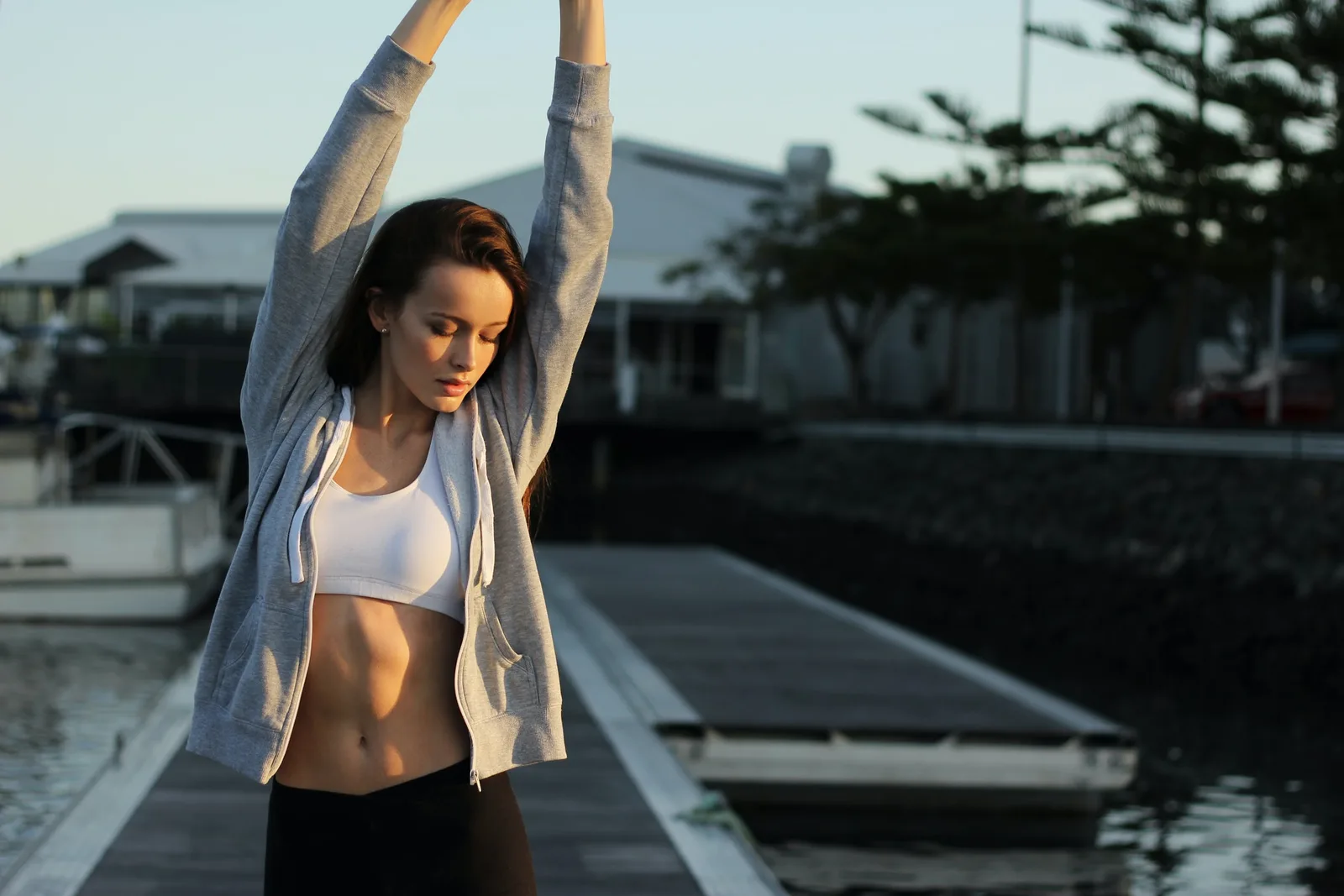 A woman in a grey hoodie and sports top stretching her arms overhead on a wooden dock in early light