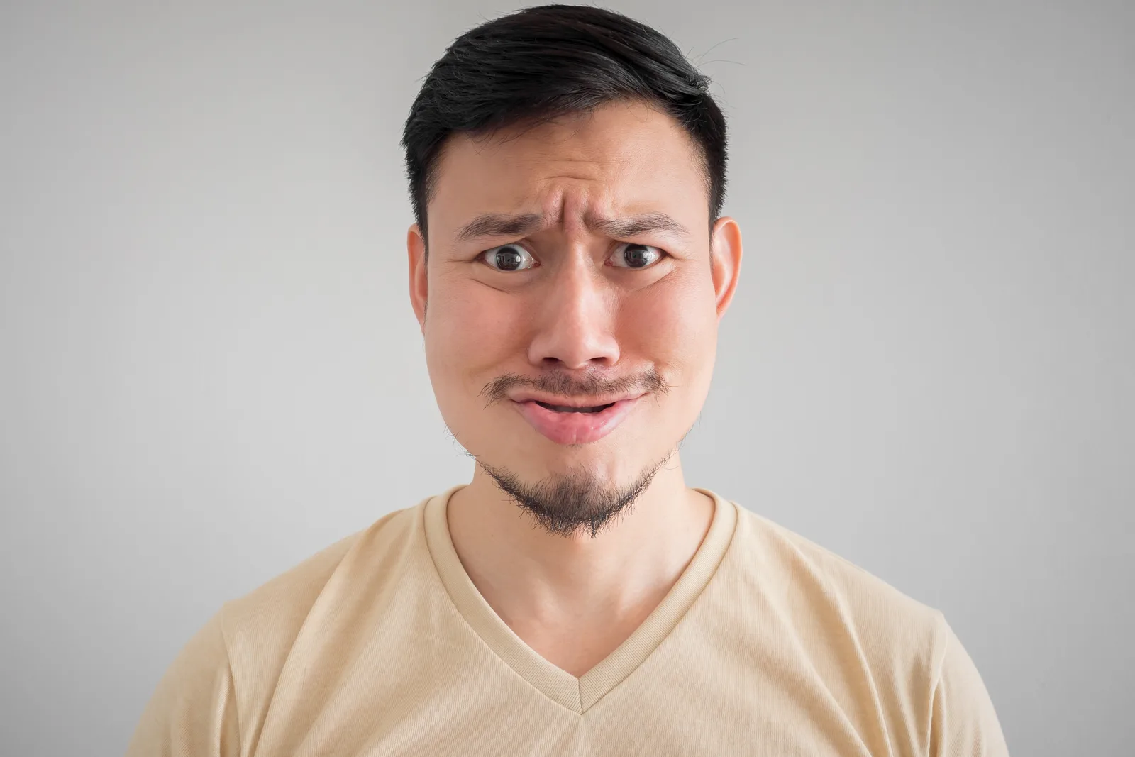 A man in a pale T-shirt with a puzzled expression, one eyebrow raised, against a grey ground